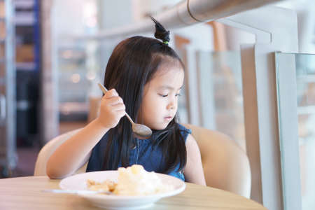 Asian children lonely or kid girl holding spoon and enjoy happy delicious eating ice cream or honey toast for sweet dessert and looking glass window on summer at kindergarten or nursery and restaurantの写真素材