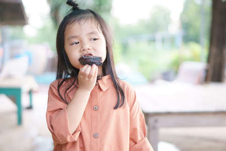 Asian child cute or kid girl enjoy and happy eating chocolate donut or doughnut and bread for delicious sweet dessert and snack at upcountry homeの写真素材