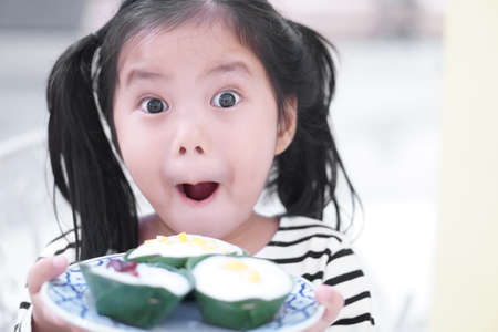Asian child cute or kid girl smiling and wow for happy and holding Thai pudding with coconut topping or Thai sweet dessert on banana leaf and white dishの写真素材