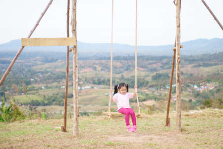 Asian child or kid girl smile playing wooden swing and happy fun or cheerful enjoy on nature holiday relax with exercise in playground meadow field or amusement public park on summer travel vacationの写真素材