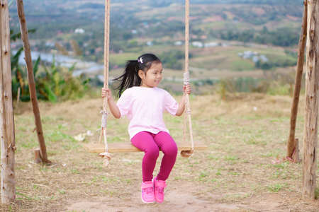Asian child or kid girl smile playing wooden swing and happy fun or cheerful enjoy on nature holiday relax with exercise in playground meadow field or amusement public park on summer travel vacationの写真素材
