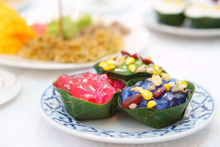 Thai pudding with coconut topping or thai sweet dessert with corn and red bean and millet in banana leaf cup and white dish on table for foodの写真素材