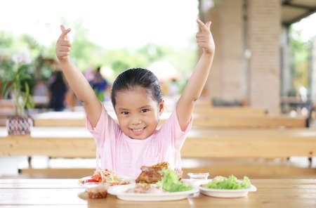 Asian child cute or kid girl enjoy eating grilled chicken with papaya salad for Thai food and appetizing with hand up on table with smiling and happy for breakfast or lunch in restaurant or food courtの写真素材