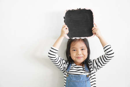 Asian child cute or kid girl happy smiling and show blank blackboard or black plate and frame on head for thinking new idea and education note at nursery school on white wall space background isolatedの写真素材