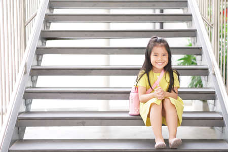 Asian child cute or kid girl smiling happy and sling water bottle or flask and sit on stairs for relax on holiday travel or waiting study to summer and thinking for new idea at nursery pre schoolの写真素材