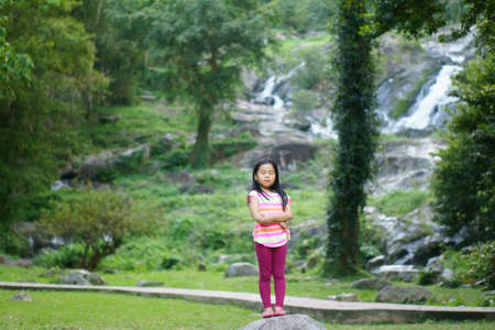 Asian child cute or kid girl happy and standing eyes closed on stone or rock with cross arm smiling on nature waterfall with green grass and tree in jungle garden for holiday relax and travel vacationの写真素材