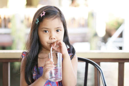 Asian child cute or kid girl drinking cool water by tube or straw in glass cup for fresh with thirsty on hot weather summer in restaurantの写真素材