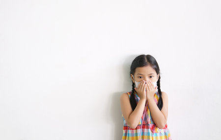 Asian child or kid girl wear N95 paper face mask and sick on nose with cold cough and protect PM2.5 dust or influenza virus and bacteria from smoke with hand close mouth on street and white backgroundの写真素材