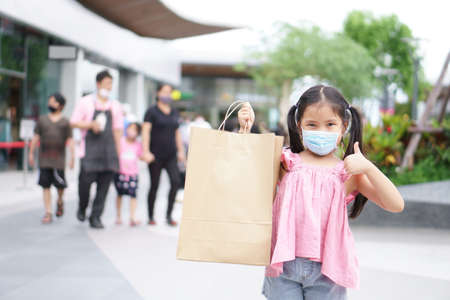 Asian child or kid girl smile wear face mask and holding empty recycle paper bag to enjoy shopping and fun with thumb up for like and good at department store or market mall to familyの写真素材