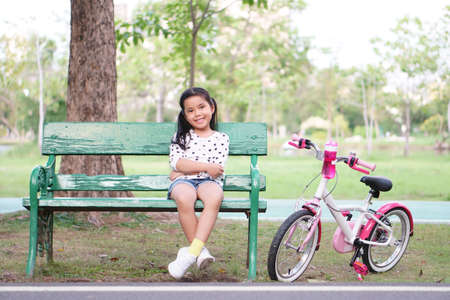 Asian child smile or kid girl sit on wood long chair with pink bicycle for relax and cycling fun with bottle water on bike in public park with green tree garden for sport exercise and healthy bikerの写真素材