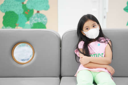 Asian child or kid girl wear N95 white face mask to close mouth nose with cold cough sick and sneeze flu to sit on sofa with social distancing for protect coronavirus or air dust at hospital or schoolの写真素材