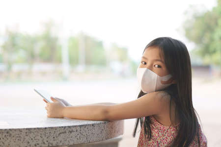 Asian child or kid girl wear N95 face mask for close mouth nose and play smartphone for online shopping and cold cough sick to protect influenza covid-19 virus or PM 2.5 dust smoke and stay home safeの写真素材
