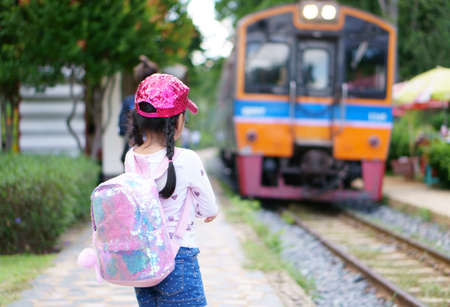 back of asian child or kid girl wait traditional train on railway or railroad in platform station with pink shoulder bag or backpack and wear cap on street for holiday travel transport and vacationの写真素材