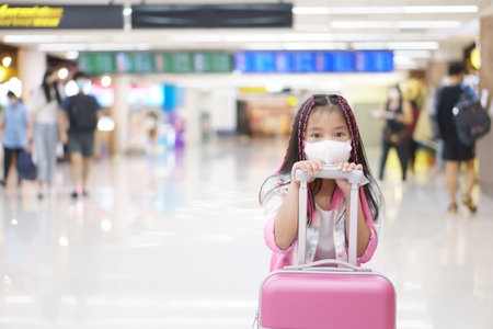 Asian child or kid girl wearing white face mask to protect from dust with dragging luggage or baggage and pink suitcase in airport for holiday travel to happy on summer vacation tripの写真素材
