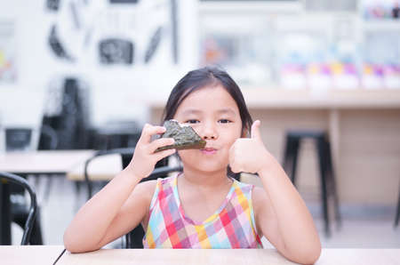 Asian child cute or kid girl enjoy eating rice ball or seaweed roll and thumbs up for japanese food like and appetizing on table and smiling to happy for breakfast or lunch in restaurant or food courtの写真素材