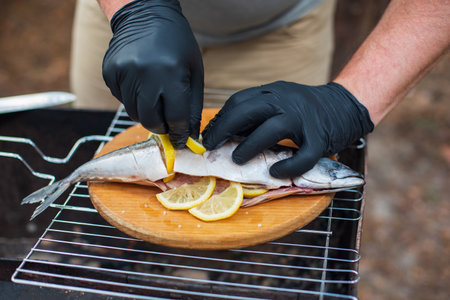 Grilled mackerel fish with lemon slices, top view. The process of cooking fish outdoors. Chef in black latex gloves preparing mackerel fish on a wooden cutting board. Tasty and fresh food, picnic, party, outdoor recreationの写真素材
