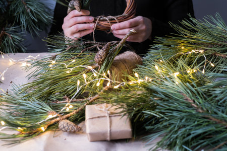 Making rustic Christmas wreath. Female hands on background of wooden table with pine tree branches,...