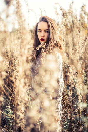 Outdoor fashion photo of young beautiful lady in autumn landscape with dry flowers. Knitted sweater, wine lipstick. Warm Autumn. Warm Springの写真素材