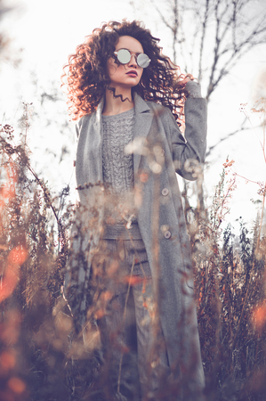 Outdoor fashion photo of young beautiful lady in autumn landscape with dry flowers. Gray coat, knitted sweater, sunglasses, wine lipstick. Fashion lookbook. Warm Autumn. Warm Springの写真素材