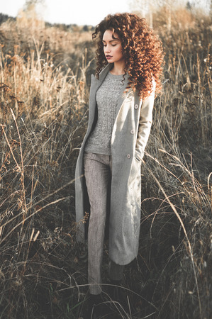 Outdoor fashion photo of young beautiful lady in autumn landscape with dry flowers. Gray coat, knitted sweater, wine lipstick. Fashion lookbook. Warm Autumn. Warm Springの写真素材