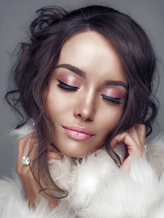 Fashion studio portrait of beautiful lady with elegant hairstyle in white fur coat. Winter beauty in luxury. Fashion fur. Beautiful woman in luxury fur coat. Fashion model posing in eco-fur coatの写真素材