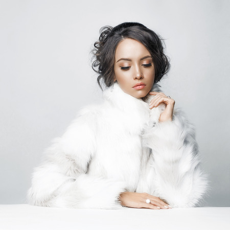 Fashion studio portrait of beautiful lady with elegant hairstyle in white fur coat. Winter beauty in luxury. Fashion fur. Beautiful woman in luxury fur coat. Fashion model posing in eco-fur coatの写真素材