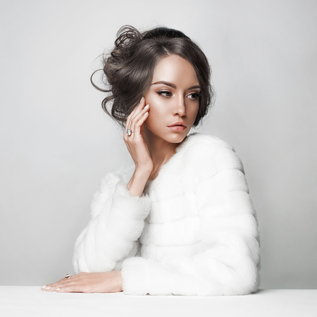 Fashion studio portrait of beautiful lady with elegant hairstyle in white fur coat. Winter beauty in luxury. Fashion fur. Beautiful woman in luxury fur coat. Fashion model posing in eco-fur coatの写真素材