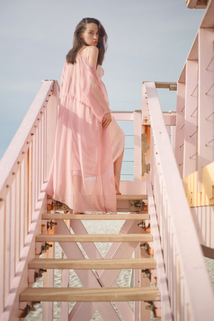 Outdoor atmospheric lifestyle portrait of beautiful pregnant woman in pink dress on pink lifeguard tower. Beautiful morning. Sunrise on the beach. Beauty and fashion. Pink aestheticの写真素材