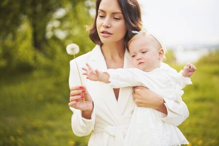 Outdoor fashion portrait of young beautiful mother and little cute daughter blowing to dandelion on...