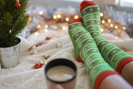 Feet in christmas socks near the Christmas tree. Woman sitting at the blanket, drinks hot beverage and relaxes warming up their feet in woollen socks. Winter and Christmas holidays conceptの写真素材