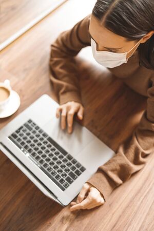 Lifestyle portrait of young lady working at notebook with mask. Woman concept for alternative office freelance. Stay home. Coronavirus. Quarantine. Online training education and freelance work. Coronavirus pandemic in the worldの写真素材