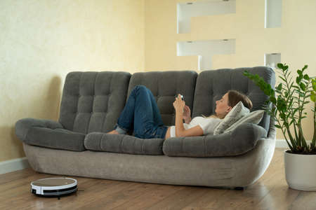 Attractive girl using smartphone while robotic vacuum cleaner cleaning houseの写真素材