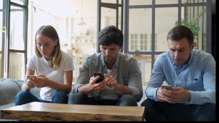 Group of three friends hanging out but ignoring each other while using their smartphones at home