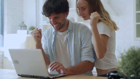 Young couple celebrating success, reading good news in email, looking at laptop screen, in kitchen at homeの写真素材