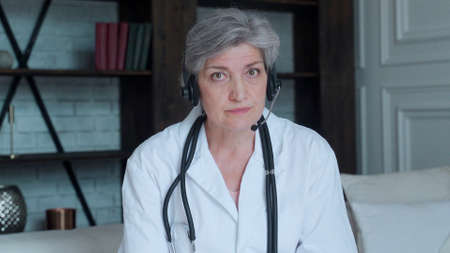 Older woman doctor therapist wearing headset video call talking to web camera consulting virtual patient online by video conference call chat.の写真素材