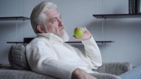 Bearded elderly man sits alone on a chair holding an Apple in his hand, thinkingの写真素材