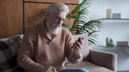 An elderly man with a gray beard is sitting on the sofa reading bad news on a smartphone