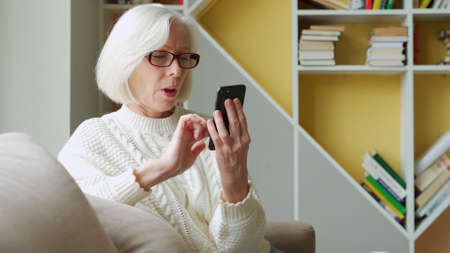 Elderly woman is sitting on the sofa, holding a smartphone, feeling very happy after receiving an...