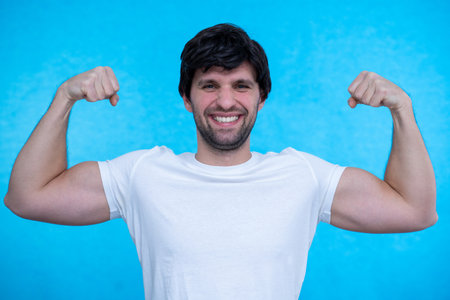 Man raises arms and shows biceps demonstrates muscles feels proud about personal achievements on a blue backgroundの写真素材
