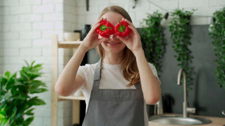 Happy young woman smiles and has fun playing with red pepper in front of her eyes in the home kitchenの写真素材