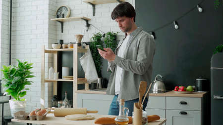 Man surfing in internet using phone standing in kitchen. Portrait of man standing in kitchen and...