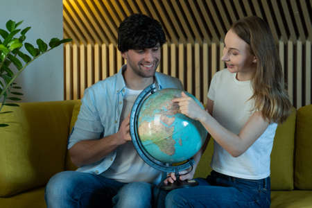 Married couple looks at the globe in the living room sitting on the sofa. Young couple choosing place for travelの写真素材