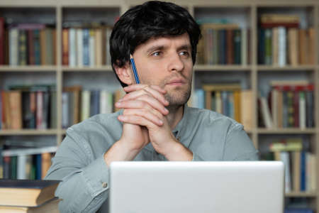 Thoughtful young man is sitting at a desk in the office, holding a pencil in his hand, looking away,...