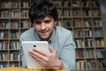 A man lying on the floor using a tablet computer at home against the background of a bookshelf