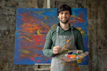 Portrait of male artist working on painting in studio. Male artist makes strokes with yellow paintの写真素材