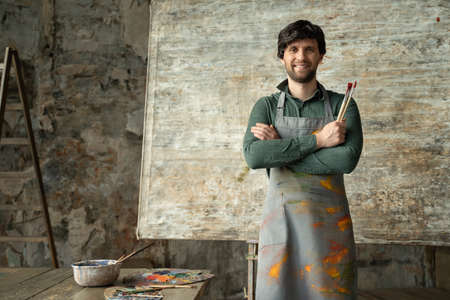 Portrait of a contented young male artist crossing his arms with a brush in his hand on a background of his new paintingの写真素材