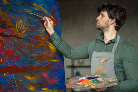 Portrait of male artist working on painting in studio. Male artist makes strokes with yellow paintの写真素材