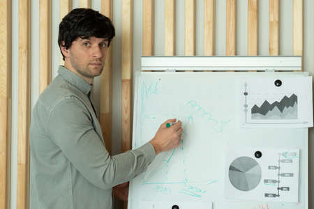 Businessman in a shirt writes on a blackboard with a marker while having a conversation with colleagues in a office, while the office staff and managers listen carefully to him in the officeの写真素材