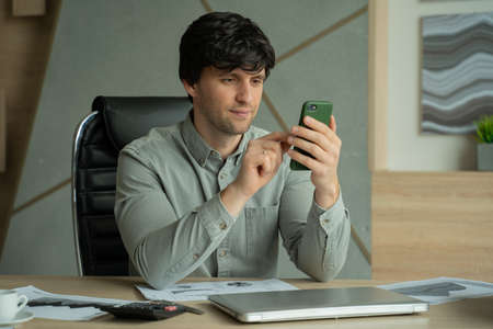 Man in a shirt sitting in an office and using a smartphone for workの写真素材