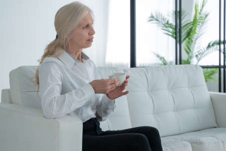 Elderly woman is sitting on the sofa, drinking coffee or tea and looking out the window